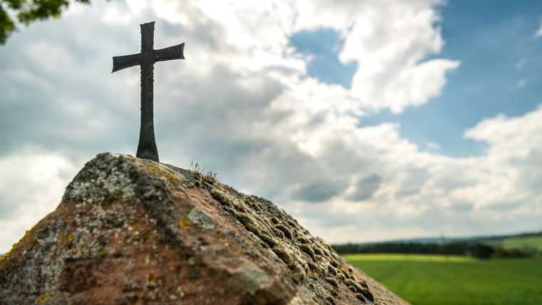 A small, black cross on a moss-covered stone in front of a cloudy sky.