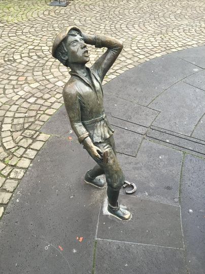Eine bronzene Statue eines Jungen, der mit einer Hand an seinem Kopf steht und nach oben schaut. Der Boden besteht aus Steinplatten und Kies.
