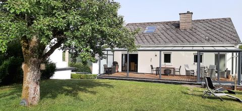 Ein modernes Haus mit einer Terrasse und einem großen Garten. Im Vordergrund steht ein Baum, der Schatten spendet.