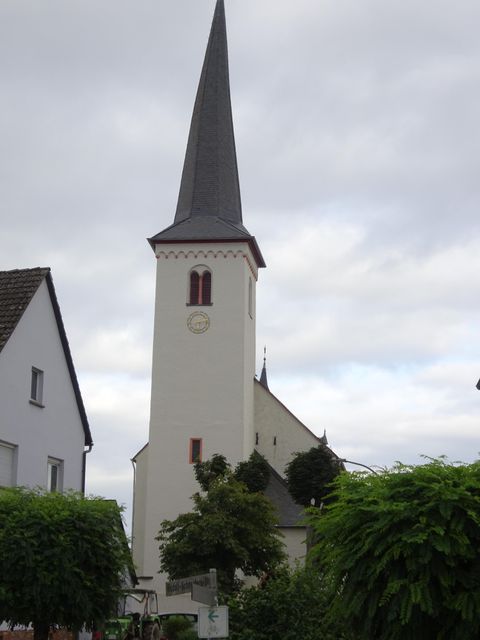 Eine Kirchengebäude mit einem hohen, spitzen Turm. Umgeben von niedrigen Häusern und grünen Pflanzen.