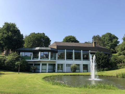 Een modern gebouw met grote ramen staat op een groene weide. Op de voorgrond bevindt zich een vijver met een fontein.