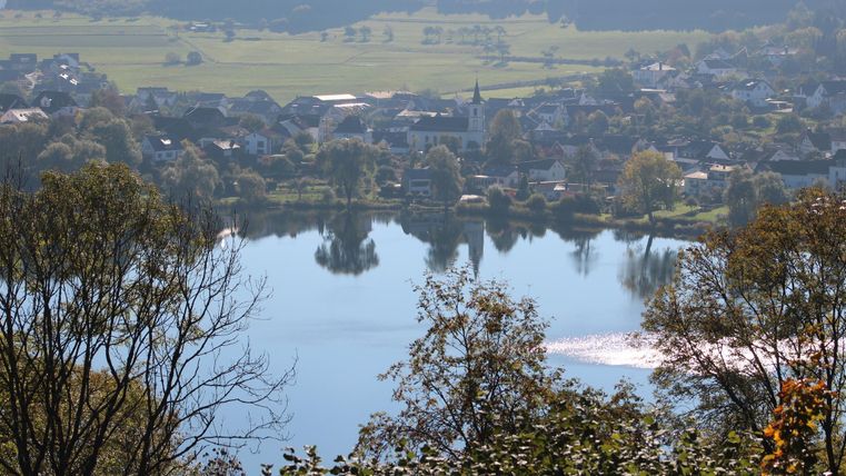 Ein ruhiger See umgeben von grüner Landschaft und kleinen Dörfern. Die spiegelnde Wasseroberfläche reflektiert die Schönheit der Natur.