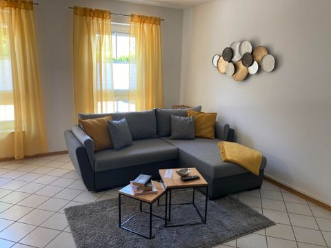 Ein gemütliches Wohnzimmer mit einer grauen Couch und gelben Vorhängen. Auf einem kleinen Tisch stehen Bücher und Dekorationsgegenstände.