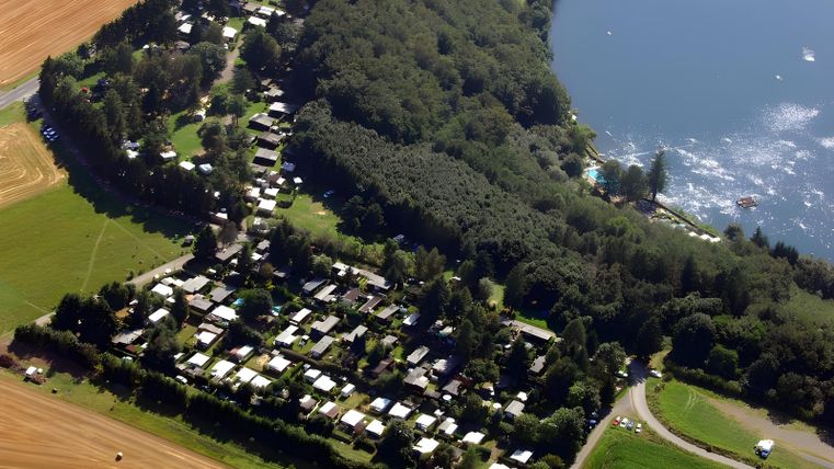Eine Luftaufnahme eines Ferienparks am Ufer eines Sees, umgeben von Bäumen und Feldern. Die Anordnung der Wohnmobile und Grundstücke ist klar erkennbar.