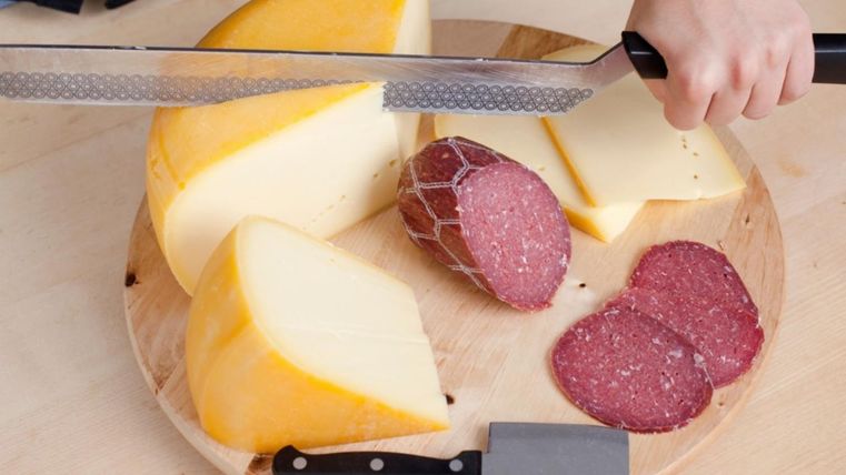 <p>Ein rundes Holzbrett, auf dem einhalbes Laib Käse liegt und mit einem großen Messer in Scheiben geschnitten wird. Daneben ein weiteres Käsemesser und eine aufgeschnittene Salami.</p>