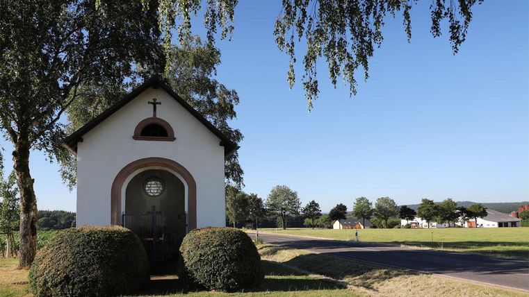 Eine kleine Kapelle mit einem Kreuz über der Tür steht neben einer Straße. Umgeben von Bäumen und Wiesen bietet sie eine ruhige Atmosphäre.