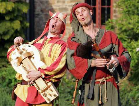 Zwei mittelalterlich gekleidete Künstler singen und spielen Musikinstrumente. Sie zeigen freudige Ausdrücke und stehen vor einer grünen Kulisse.