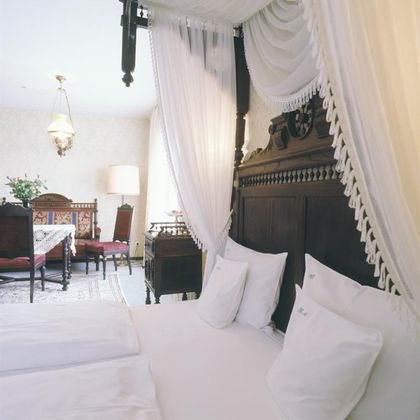 Ein gemütliches Schlafzimmer mit einem großen, dekorativen Bett und eleganter Bettwäsche. Im Hintergrund befindet sich ein stilvoll eingerichteter Wohnbereich mit Tisch und Stühlen.