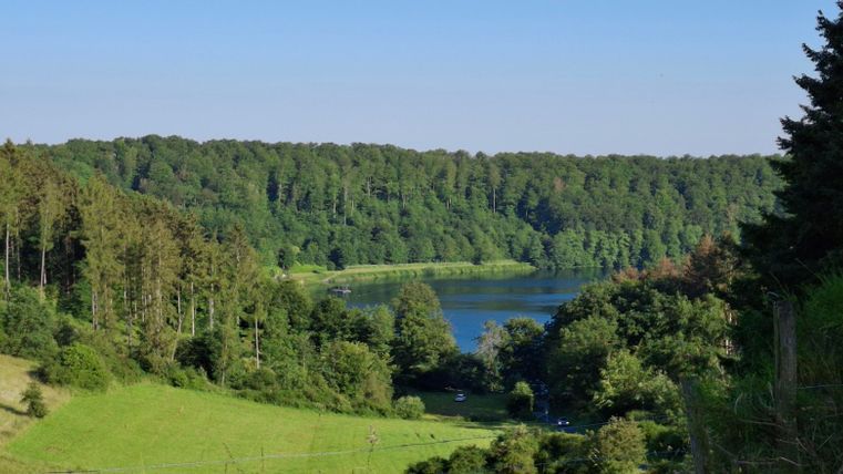 A picturesque landscape with a tranquil lake and surrounding forests. The sky is clear, and the vegetation is lush and green.