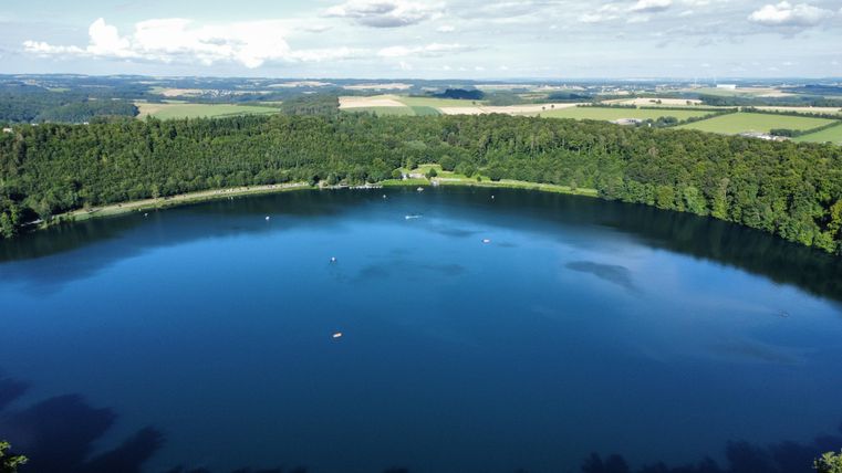 Ein ruhiger See umgeben von grünen Wäldern und Wiesen. Die wasserfläche spiegelt den klaren Himmel wider.
