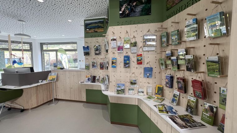Ein moderner Informationsraum mit einer Wand voller Broschüren über Wanderrouten. Der Raum hat helle Farben und viel Tageslicht.