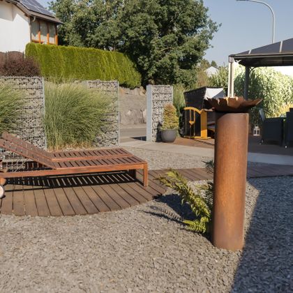 A modern garden landscape featuring a wooden lounge chair on a round wooden deck. Tall grasses and a gazebo can be seen in the background.
