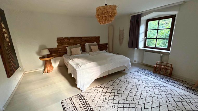 Ein modernes Schlafzimmer mit einem großen Bett und dekorativen Kissen. Der Raum hat natürliche Materialien und ein Fenster mit Blick ins Freie.