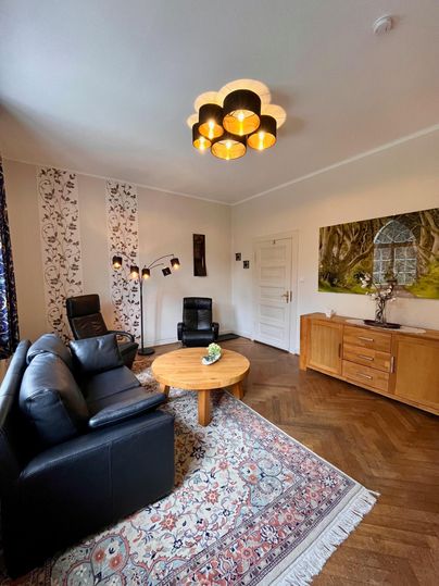 Ein stilvolles Wohnzimmer mit einem runden Tisch und einer modernen Lampe. Die Einrichtung umfasst eine schwarze Couch und zwei bequeme Sessel auf einem schönen Teppich.