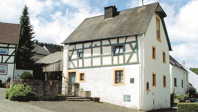 Ein traditionelles Fachwerkhaus mit weißer Fassade und orangefarbenen Akzenten. Die Umgebung ist ländlich mit einigen Bäumen und einem gepflasterten Weg.