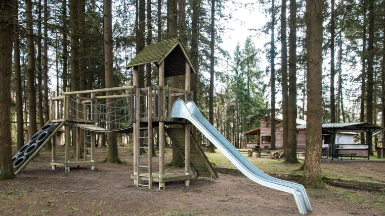 Ein Spielplatz im Wald mit einer Holz-Spielift und einer Rutsche. Umgeben von hohen Bäumen gibt es auch einen Bereich mit Picknicktischen.