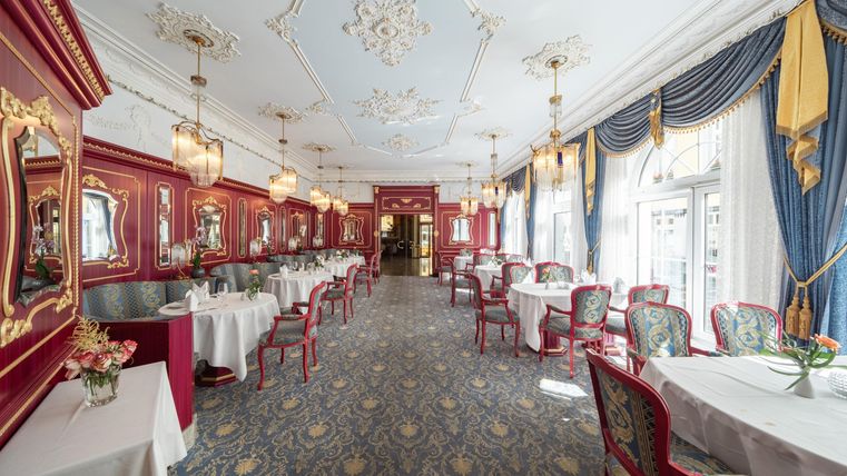 Ein eleganter Speisesaal mit roten Wänden und stilvollen Möbeln. Große Fenster lassen viel Licht herein und sorgen für eine einladende Atmosphäre.