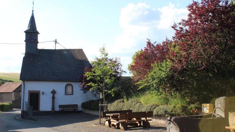Eine kleine Kirche mit einem schlanken Turm steht an einer ruhigen Straße. Umgeben von Bäumen und Grünfläche strahlt der Ort eine friedliche Atmosphäre aus.