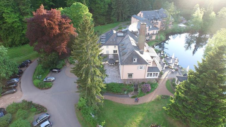 Ein malerisches Anwesen mit einem gepflegten Garten und einem kleinen See. Umgeben von Bäumen gibt es eine Zufahrt mit parkenden Autos.
