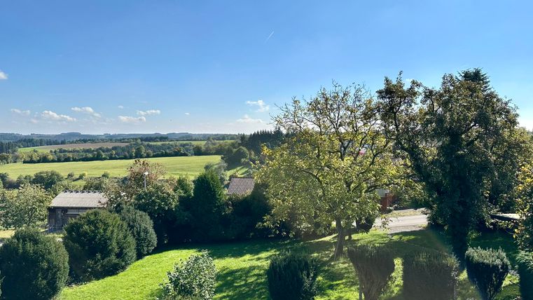 Eine weitläufige Landschaft mit grünen Wiesen und Bäumen unter einem klaren blaube Himmel. Im Hintergrund sind sanfte Hügel und einige Gebäude zu sehen.