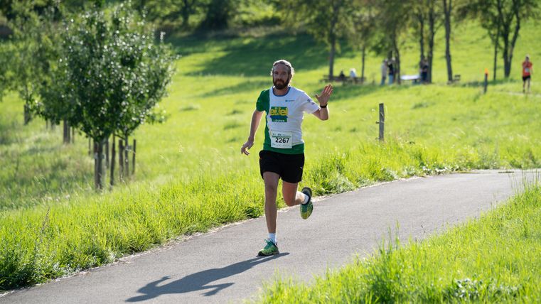 Ein Läufer in einem weißen T-Shirt joggt auf einem Weg in einer grünen Landschaft. Im Hintergrund sind weitere Personen und Bäume zu sehen.