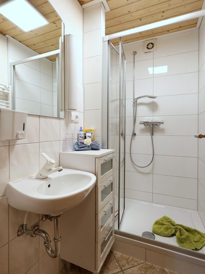 Ein modernes Badezimmer mit einer Dusche und einem Waschbecken. Die Wände sind hell gefliest, und ein Handtuch liegt auf dem Boden.