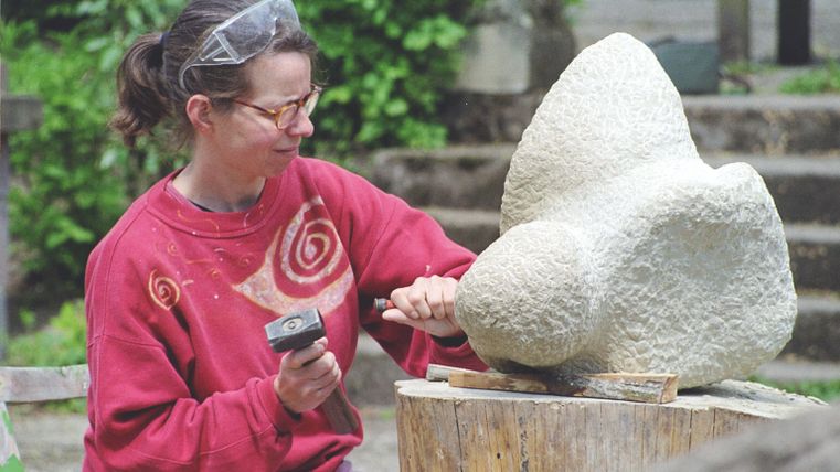 Eine Künstlerin bearbeitet einen Steinblock mit einem Hammer und Meißel. Im Hintergrund sind grüne Pflanzen und eine entspannte Umgebung.