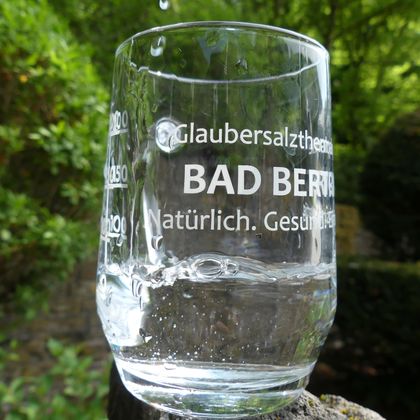 Ein Glas mit Wasser steht auf einem Stein. Auf dem Glas steht der Text "Glaubersalztherme BAD BERG" sowie "Natürlich. Gesund."