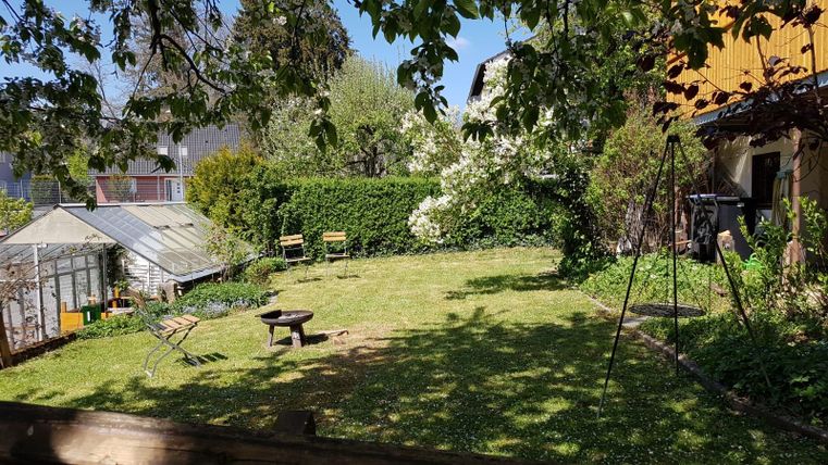 Ein grüner Garten mit einer Rasenfläche und blühenden Bäumen. Im Hintergrund befinden sich eine Gartenhütte und zwei Bänke.