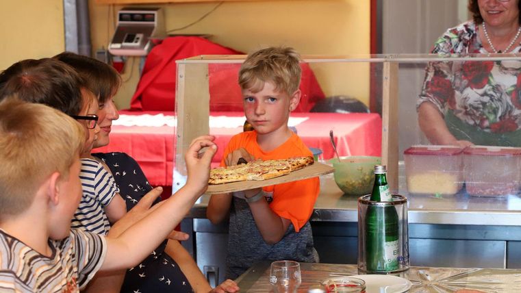 Eine Gruppe von Kindern sitzt an einem Tisch und schaut auf einen Jungen, der eine Pizza auf einem Teller hält. Im Hintergrund ist eine Frau zu sehen, die in der Küche arbeitet.