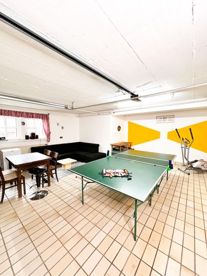 Ein heller Raum mit einem Tischtennistisch und mehreren Sitzgelegenheiten. An der Wand sind gelbe Akzente und ein Fenster mit Vorhängen zu sehen.