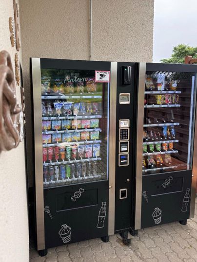 Zwei Getränke- und Snackautomaten stehen nebeneinander an einer Wand. Sie bieten eine Vielzahl von Snacks und Erfrischungen an.