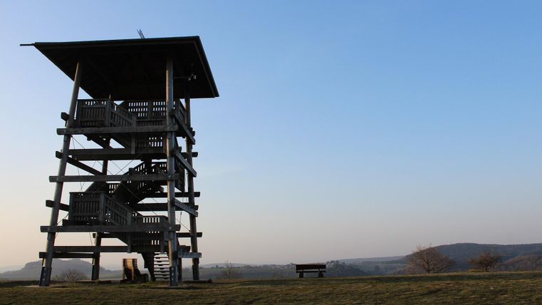 Ein Aussichtsturm auf einem Hügel mit klarem Himmel im Hintergrund. Der Boden ist grün und es gibt eine Bank in der Nähe.