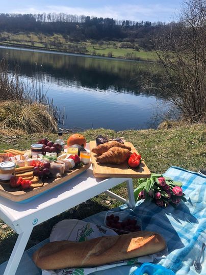 Ein schön gedeckter Picknicktisch am Ufer eines ruhigen Sees. Auf dem Tisch sind verschiedene Speisen, Brot und Blumen zu sehen.