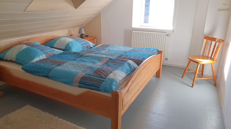 Ein helles Schlafzimmer mit einem Holzbett und bunten Bettdecken. Neben dem Bett steht ein Holzstuhl und ein Fenster bringt Tageslicht herein.