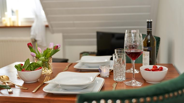 Ein elegant gedeckter Tisch mit zwei Tellern, Gläsern und einer Flasche Wein. Frische Blumen und Obst runden die gemütliche Atmosphäre ab.