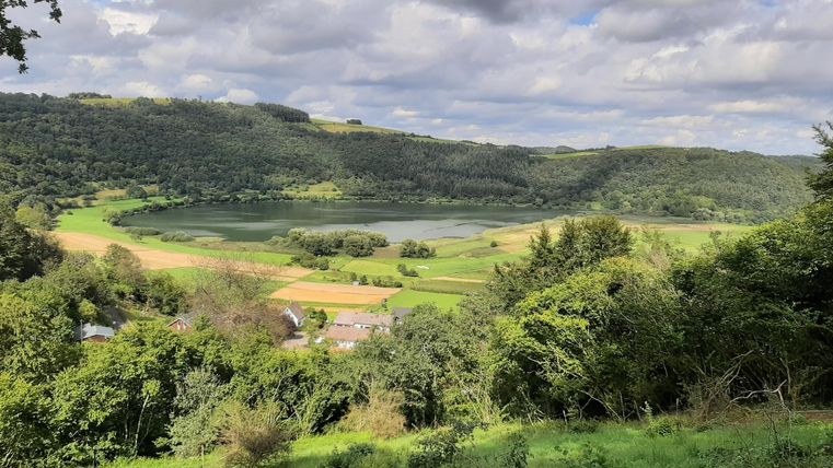 Eine malerische Landschaft mit einem ruhigen See und sanften Hügeln. Der Himmel ist bewölkt und die Umgebung ist grün und üppig.