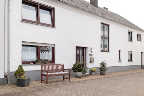 Ein weißes Haus mit einer modernen Fassade und großen Fenstern. Vor dem Haus steht eine Holzbank und einige Pflanzen in Töpfen.