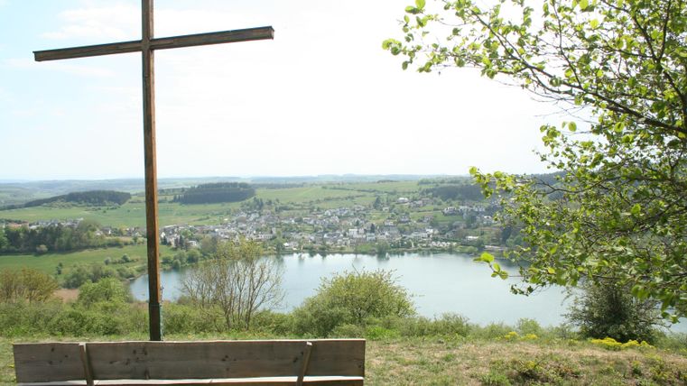 Ein Aussichtspunkt mit einem großen Holzkreuz und einer Bank. Im Hintergrund sieht man einen malerischen See und das umliegende grüne Land.