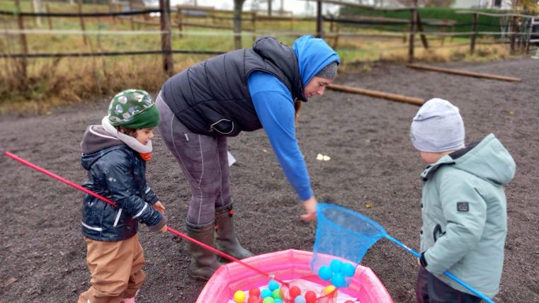 Eine Frau spielt mit zwei Kindern im Freien. Sie fangen bunte Kugeln aus einem pinken Becken.
