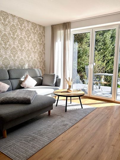 Ein gemütliches Wohnzimmer mit einem grauen Sofa und einem kleinen Tisch. Große Fenster lassen viel Licht herein und bieten einen Blick ins Grüne.
