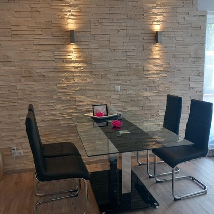 A modern dining area with a glass table and black chairs. The walls are decorated with stone slabs, and two wall sconces create a cozy atmosphere.