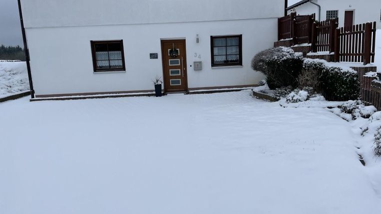 Ein Haus im Schnee mit einem klaren, weißen Hof. Einige Pflanzen sind sichtbar, und es gibt einen Zaun im Hintergrund.