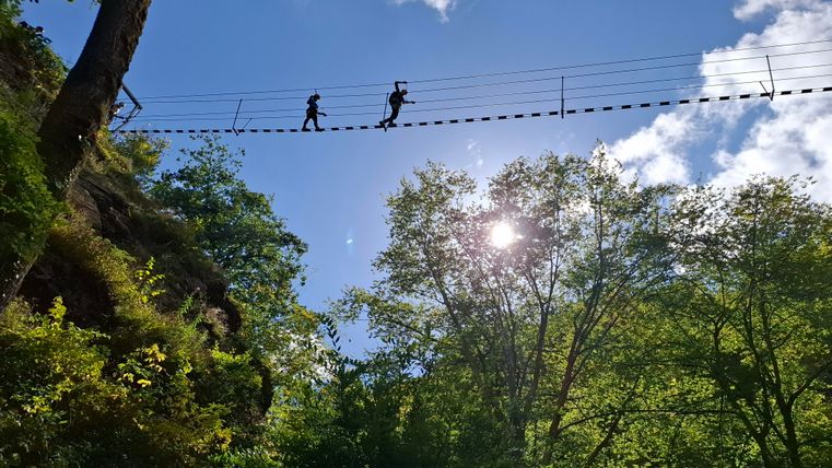 Eine schmale Hängebrücke überquert einen Wald mit dichten Bäumen. Zwei Personen gehen auf der Brücke, während die Sonne durch die Wolken scheint.