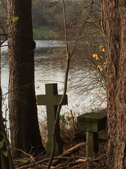 Ein ruhiger Ort am Wasser, umgeben von Bäumen. Ein Kreuz und eine Bank sind sichtbar, was die friedliche Atmosphäre verstärkt.
