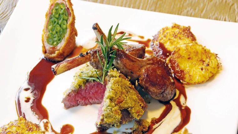 Ein kunstvolles Gericht mit Lammkoteletts, umgeben von knusprigen Beilagen und einer Sauce. Das Essen ist dekorativ angerichtet und zeigt frische Kräuter.