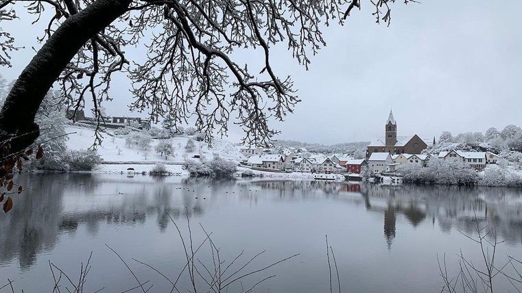 Ein winterliches Landschaftsbild mit schneebedeckten Bäumen und einem ruhigen See. Im Hintergrund sind einige Häuser und ein Kirchturm sichtbar.