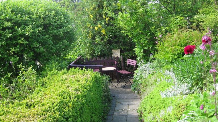 Ein schöner Gartenweg mit dichtem grünen Gestrüpp. Auf einer kleinen Terrasse stehen ein Tisch und zwei Stühle.
