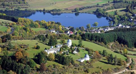 A peaceful landscape with a clear lake and colorful trees. In the background, gentle hills and small buildings can be seen.