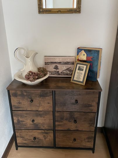 Ein eleganter Holzschrank mit sechs Schubladen steht an der Wand. Darauf befinden sich eine Kanne, ein Bild, ein gerahmtes Dokument und einige Deko-Elemente.